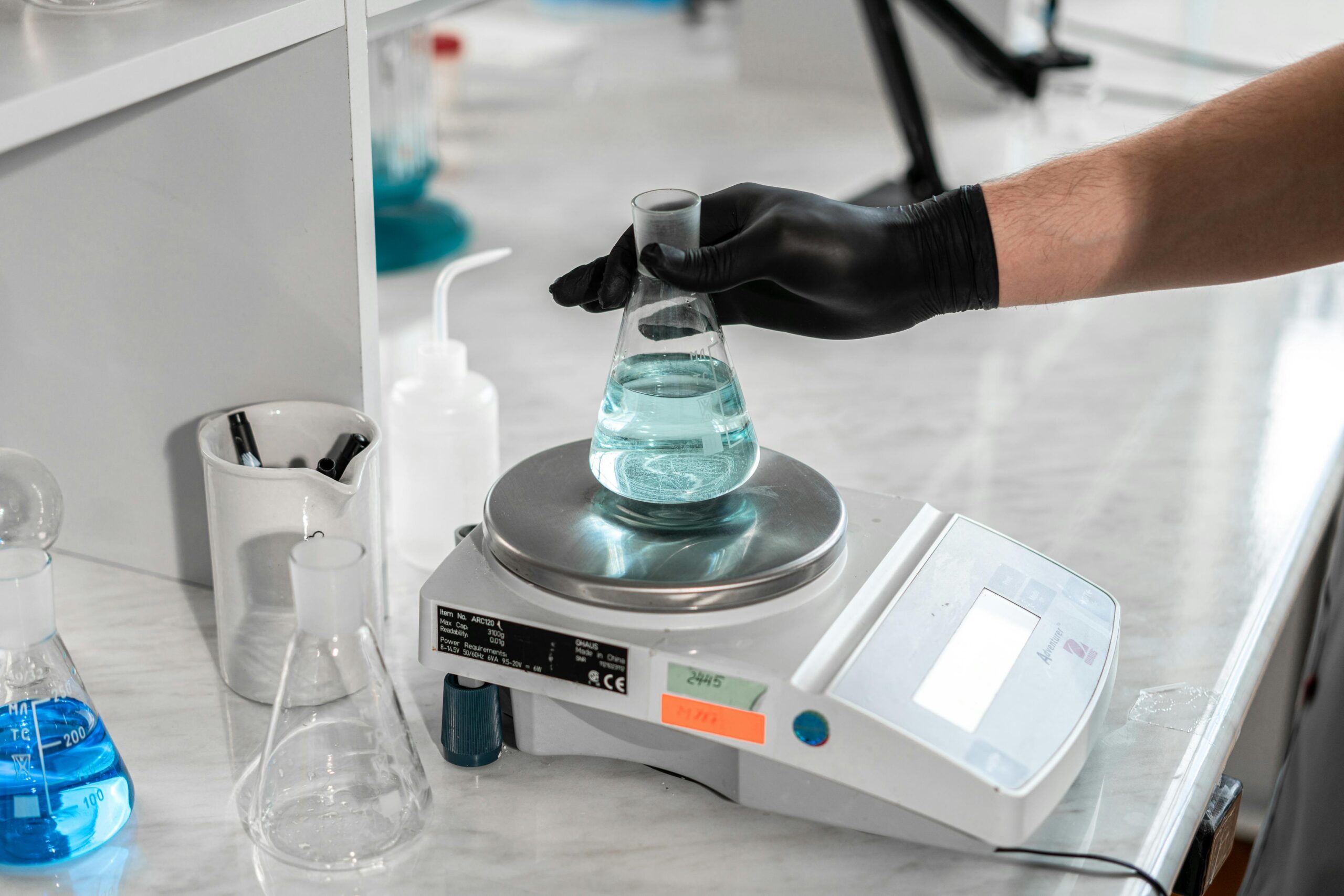 A scientist handling a liquid-filled flask on a laboratory scale for precise measurement.